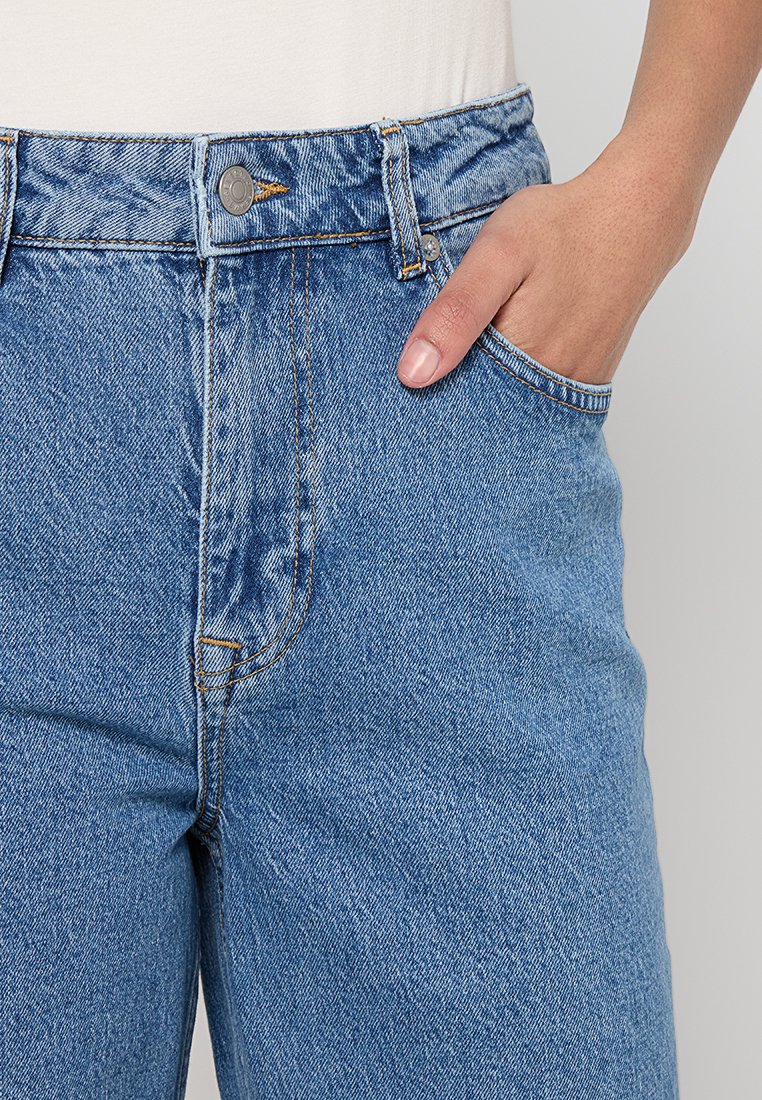 Jeans z wysokim stanem w niebieskim kolorze, z lekko wyblakłą teksturą, z przednią kieszenią i kontrastującymi żółtymi szwami.