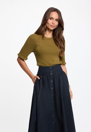 Kvinde med langt brunt hår iført en olivengrøn tekstureret sweater og et højt mørkeblåt nederdel med knapper, stående med den ene hånd i lommen.