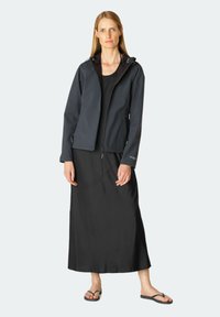 Zwarte waterdichte jas met capuchon, slimme pasvorm, ritssluiting en zijzakken, gedragen over een lange zwarte jurk en slippers.