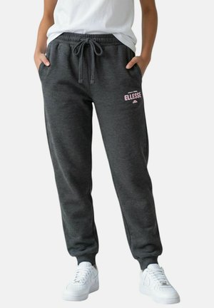 Personne portant un pantalon de survêtement gris foncé à cordon avec le logo "ellesse" sur la cuisse, des baskets blanches et un t-shirt blanc, les mains dans les poches.