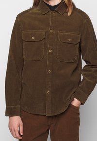 Camisa de corduroy marrón con collar abotonado, dos bolsillos en el pecho y cierre frontal de botones. Presenta una textura acanalada y un dobladillo curvado.