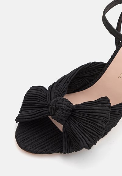 Zapato de tela negra con un diseño plisado texturizado, que presenta un prominente lazo anudado en la parte frontal y un forro interior beige.