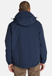 Una persona con el pelo corto que lleva una chaqueta con capucha de color azul oscuro, vista desde atrás, apoyada contra un fondo blanco liso.