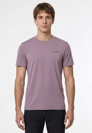 Giovane uomo che indossa una t-shirt a maniche corte mauve di Vaude e pantaloni neri, in piedi rivolto verso la fotocamera con un'espressione neutra.