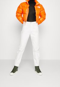 Jasnopomarańczowa kurtka puchowa, czarny golf, białe dżinsy i oliwkowozielone sneakersy za kostkę. Kurtka ma skrócony krój i logo.