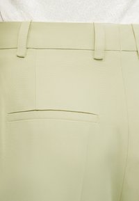 Φωτεινό μπεζ παντελόνι με θηλιές για ζώνη και μια πίσω τσέπη με φάσα, φορεμένο με λευκό υφασμάτινο τοπ.