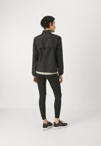 Persona in piedi, con giacca nera, leggings neri e sneaker nere con dettagli viola, su uno sfondo chiaro e semplice.