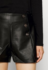 Černé kožené šortky s vysokým pasem, které mají hladkou texturu, pět knoflíkových detailů a boční švy. Kombinují se s černým topem s dlouhým rukávem.