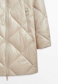 Veste matelassée beige avec motif à carreaux diagonaux, fermeture éclair frontale et une poche zippée verticale sur la poitrine.