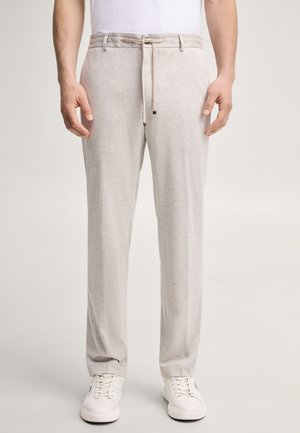 Uomo che indossa pantaloni da casa grigio chiaro con coulisse e sneakers bianche, in piedi contro uno sfondo chiaro e semplice, visibili busto e gambe.