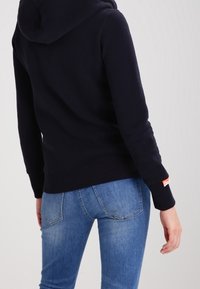 Mörkblå hoodie med känguruficka, ribbade muddarna och nederkant, justerbar dragsko på huvan och orange etikett detalj på ärmen.