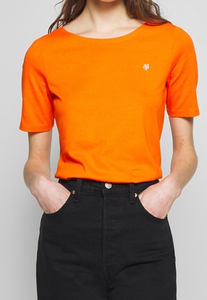 Femme portant un t-shirt orange vif à manches courtes avec un petit logo blanc sur la poitrine, les mains dans les poches d’un pantalon noir taille haute.