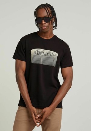 T-shirt en coton noir avec un logo G-Star texturé et ondulé en blanc et gris. Le mannequin porte des lunettes de soleil, les mains jointes devant.