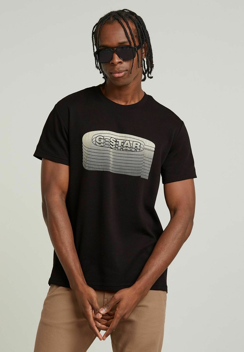 Camiseta de algodón negra con un logo de G-Star texturizado y ondulado en blanco y gris. El modelo lleva gafas de sol y las manos juntas por delante.