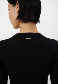 Svart ribbestrikket genser med en klassisk rund hals, med detalj av gulllogo bak. Glatt tekstur, tettsittende design.
