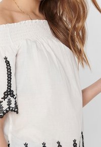 Haut blanc à épaules dénudées avec broderie noire le long des manches et de l'ourlet, présentant une coupe décontractée et un tissu doux et léger.