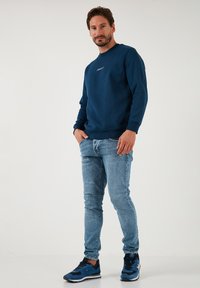 Tumesinine sweatshirt ümarate kaelustega ja valge logo, koos helesiniste kitsaste teksade ja tumesiniste sporditossudega, lihtsal taustal.