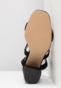 Sandale noire à talon haut avec une tige en tissu noir texturé, design à bout ouvert, et une semelle en caoutchouc marron clair présentant des rainures horizontales.