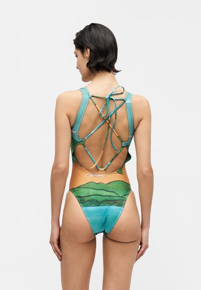 Γυναίκα με κοντά σκούρα μαλλιά που φοράει ολόσωμο μαγιό με σχέδιο χωρίς πλάτη και πολύχρωμο τοπία, φαίνεται από πίσω.