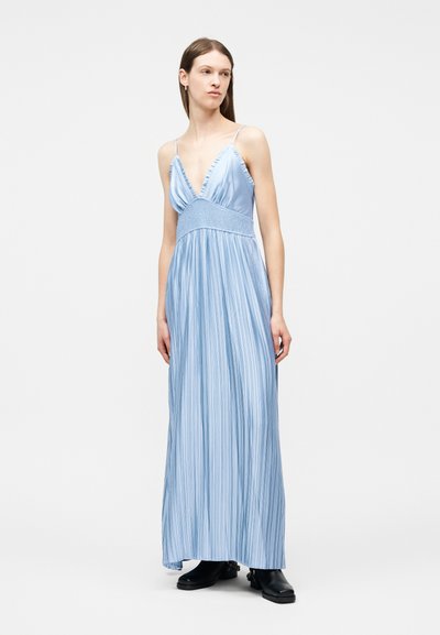 Γυναίκα που φοράει αμάνικο μακρύ φόρεμα σε ανοιχτό γαλάζιο χρώμα με πιέτες και βαθύ V-λαιμόκοψη και μαύρα μποτάκια αστραγάλου, στέκεται μπροστά σε λευκό φόντο.