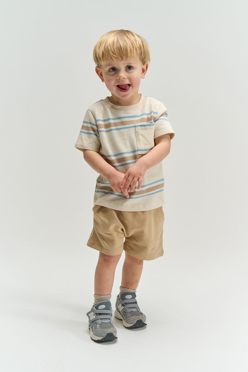 Jeune garçon blond portant un t-shirt rayé beige, un short beige, des baskets gris, debout avec les mains jointes et la langue légèrement sortie.