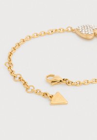 Βραχιόλι σε χρυσό τόνο με σχεδιασμό αλυσίδας, που διαθέτει ένα μενταγιόν σε σχήμα καρδιάς διακοσμημένο με στράς και μια τριγωνική πλακέτα με χαραγμένο το "GUESS".