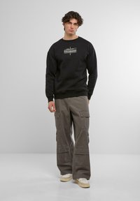Sudadera negra con logo en el pecho, combinada con pantalones cargo grises de corte holgado que cuentan con bolsillos. El modelo lleva zapatillas color beige.