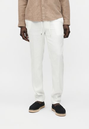 Férfi fehér zsinóros nadrágban, fekete, szőtt talpú papucsban, és világosbarna, hosszú ujjú, gombos ingben.