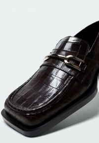 Mørkebrun læderloafer med krokodillemønster og sølvmetalspænde, firkantet tå og lav hæl på hvid baggrund.