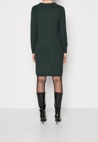 Abito lavorato a maglia verde scuro con maniche lunghe, silhouette aderente e fondo a coste. Abbinato a stivali neri con tacco al ginocchio.