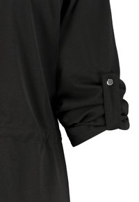 Vêtement noir à manches longues avec revers de manche retroussé maintenu par une patte en tissu et un bouton-pression, présentant une texture de tissu lisse et légèrement froncée.