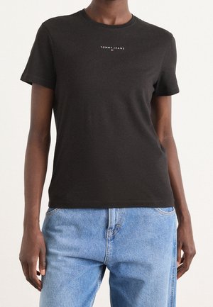 Persoon met een zwarte Tommy Jeans T-shirt met ronde hals gecombineerd met lichtblauwe spijkerbroek, staande tegen een effen achtergrond.