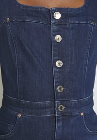 Jeansoverall i mörkblått, med silverfärgade tryckknappar, en figurnära design och ett bälte i midjan samt fickor på sidorna.