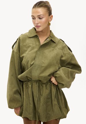 Veste oversize vert olive avec fermeture éclair, col roulé et taille froncée. Tissu doux avec une légère texture et manches volumineuses.