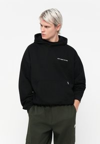 Zwarte cropped hoodie met capuchon en vozakken. Heeft een witte geborduurde tekst "VERSPEEL DE CULTUUR NIET" aan de linkerkant. Zacht materiaal.