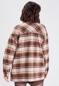 Personne aux cheveux ondulés mi-longs portant une chemise à manches longues à carreaux marron, crème et orange, vue de dos sur un fond blanc.