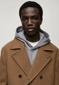 Bruine wollen jas met een dubbelzijdig ontwerp, grote revers en zwarte knopen, gedragen over een grijze hoodie met een trekkoord.