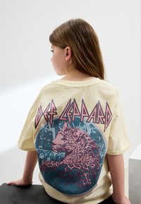 Übergroßes cremefarbenes T-Shirt mit einer Grafik eines Leoparden und dem Schriftzug "Def Leppard" in Rot, vor einem blauen kreisförmigen Hintergrund und mit Retro-Designelementen.