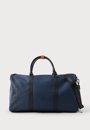 Temno modra torba duffel iz trpežne tkanine, s črnimi usnjenimi poudarki, dvema zgornjima ročajema in odstranljivim naramnim trakom z logotipom.