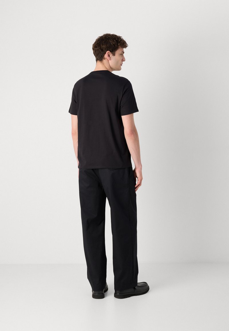 Camiseta negra de manga corta con cuello redondo, combinada con pantalones holgados negros y calzado negro, vista desde la parte posterior.