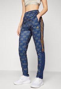 Siniset urheiluhousut, joissa on palmukuviot, kultaiset sivuraidat, joustava vyötärönauha ja taskut; yhdistetty valkoisiin lenkkareihin.