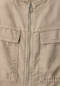 Chaqueta de tela beige, vista frontal con cremallera metálica y dos bolsillos con solapa, costuras detalladas alrededor de los bolsillos y el área de la cremallera.