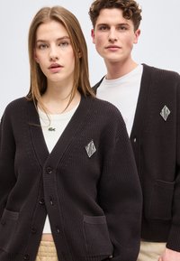 Νεαρή γυναίκα και άνδρας στέκονται δίπλα δίπλα φορώντας μαύρα πλεκτά ζακέτες πάνω από λευκά πουκάμισα, αντικρίζοντας μπροστά μπροστά σε απλό ανοιχτό φόντο.