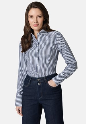 Kobieta o długich brązowych włosach, ubrana w niebiesko-białą koszulę w pionowe paski na guziki i ciemnoniebieskie dżinsy, z jedną ręką w kieszeni.