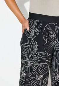 Zwarte broek met een bloemenpatroon in het wit, met een brede tailleband en zijzakken. Glad stofoppervlak met omlijnde bloemendetails.