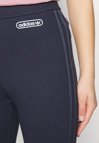 Granatowe prążkowane legginsy z białym logo Adidas, z trzema ciemnymi pionowymi paskami po bokach i elastycznym pasem.