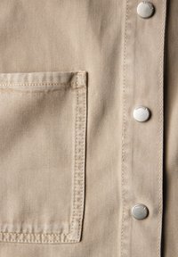 Tissu en denim beige avec une poche à rabat, des boutons-pression en argent et une texture diagonale subtile. Gros plan sur le détail du vêtement.