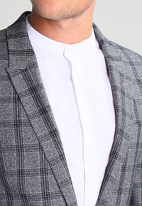 Mężczyzna noszący szary, w kratę marynarkę na białej koszuli z mandaryńskim kołnierzykiem, widoczny od szyi do górnej części klatki piersiowej.