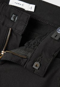 Pantalon noir avec une braguette à zip à dents dorées, fermeture par bouton pression, et une étiquette en tissu indiquant les informations de taille.