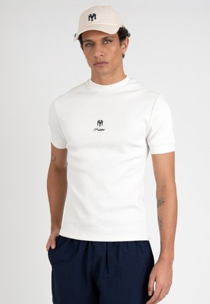 Mand iført en hvid tætsiddende t-shirt med sort logo og tekst, beige kasket med logo og mørkeblå bukser, stående mod en ensfarvet baggrund.